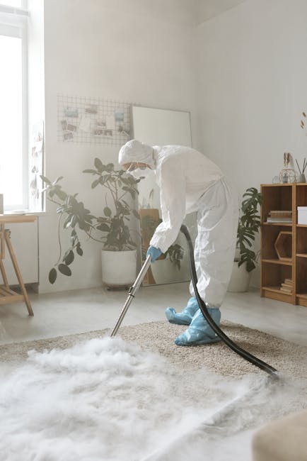 A man with curly dark hair and a beard, dressed in an orange long-sleeve shirt and gray trousers, is bent over in a living room, carefully adjusting a beige textured carpet with his right hand. The ro
