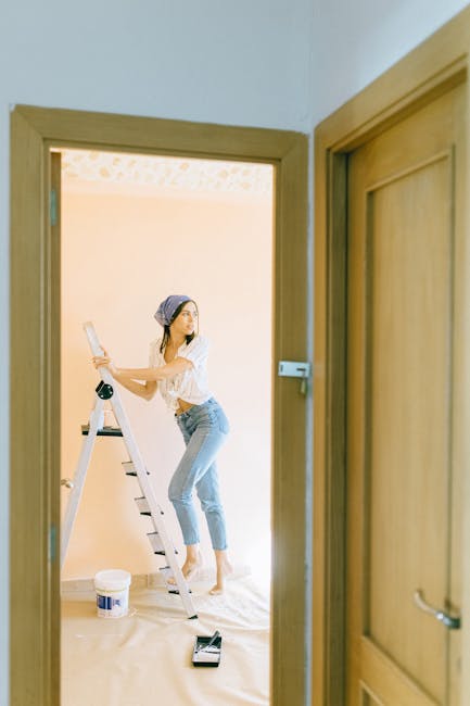 A young woman standing on a white step ladder performing domestic cleaning in a well-lit room with cream-colored walls. She is wearing a light-colored headscarf, white top, and light blue jeans, and i