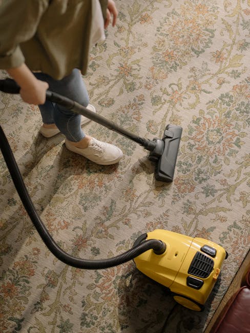 Close-up view of a person vacuuming a beige, textured area rug in a residential living space with natural light illuminating the room. The vacuum cleaner is red with a blue logo and is placed on the r