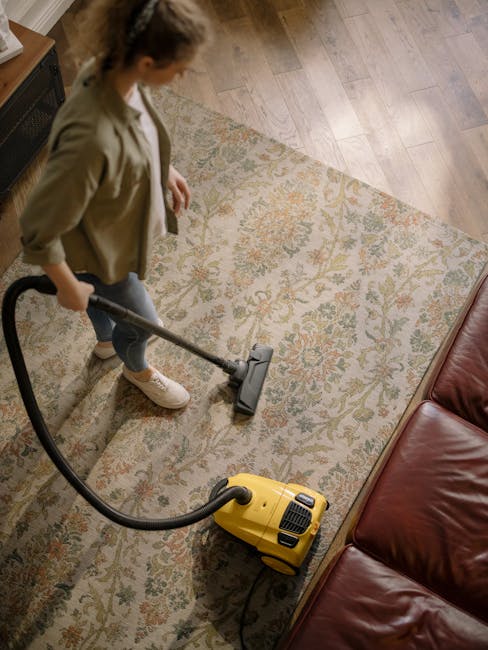 A man with curly black hair and a beard is crouched down on a wooden floor, adjusting a beige carpet with a textured pattern. He is wearing an orange long-sleeve shirt and light gray pants, with red s